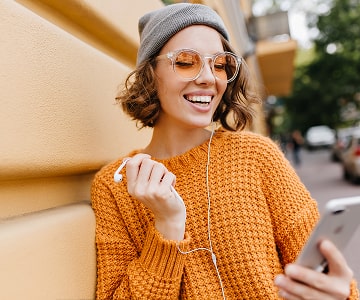 Smiling young woman in orange sweater studying online courses on smatphone